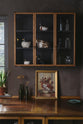 A quiet corner of a warm and inviting Haberdasher’s kitchen features a long sink run with a copper worktop, and glazed wall cupboards. Adorned with a luscious green potted plant, vintage items, artisan ceramics, and a golden framed floral painting, this kitchen creates a lovely exhibitionist space that is both beautiful and practical.