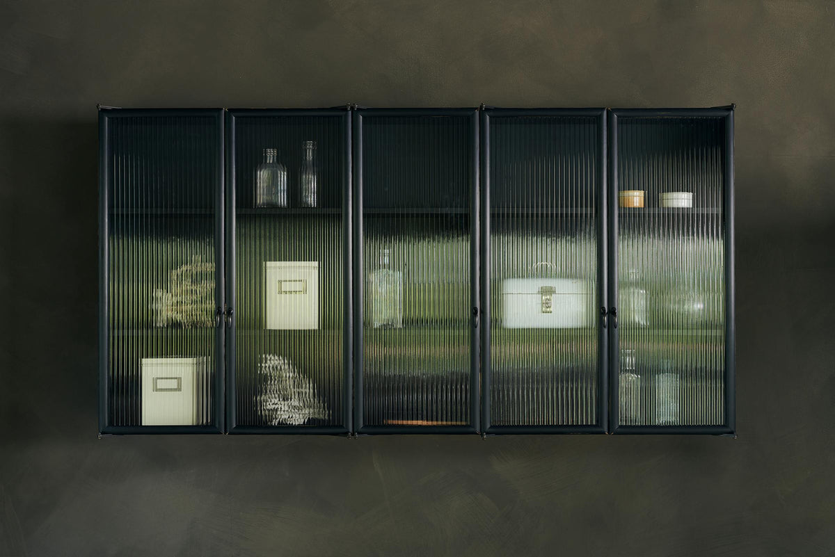 A bobbin black Haberdasher’s wall cupboard with reeded glass hangs on an olive limewash wall. Adorned with vintage white tin cases, empty glasses, and coral decorations, this space creates a lovely exhibitionist area that is both beautiful and practical.