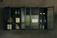 A bobbin black Haberdasher’s wall cupboard with reeded glass hangs on an olive limewash wall. Adorned with vintage white tin cases, empty glasses, and coral decorations, this space creates a lovely exhibitionist area that is both beautiful and practical.