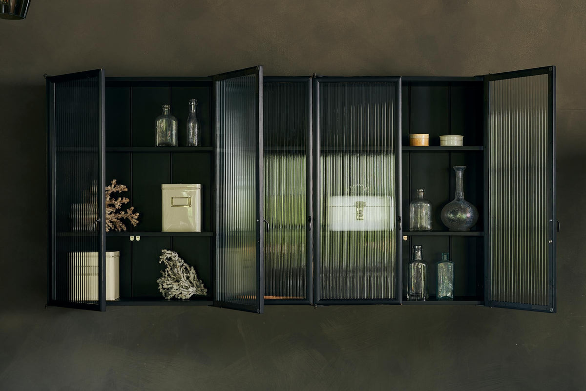 A bobbin black Haberdasher’s wall cupboard with reeded glass hangs on an olive limewash wall. Adorned with vintage white tin cases, empty glasses, and coral decorations, this space creates a lovely exhibitionist area that is both beautiful and practical.