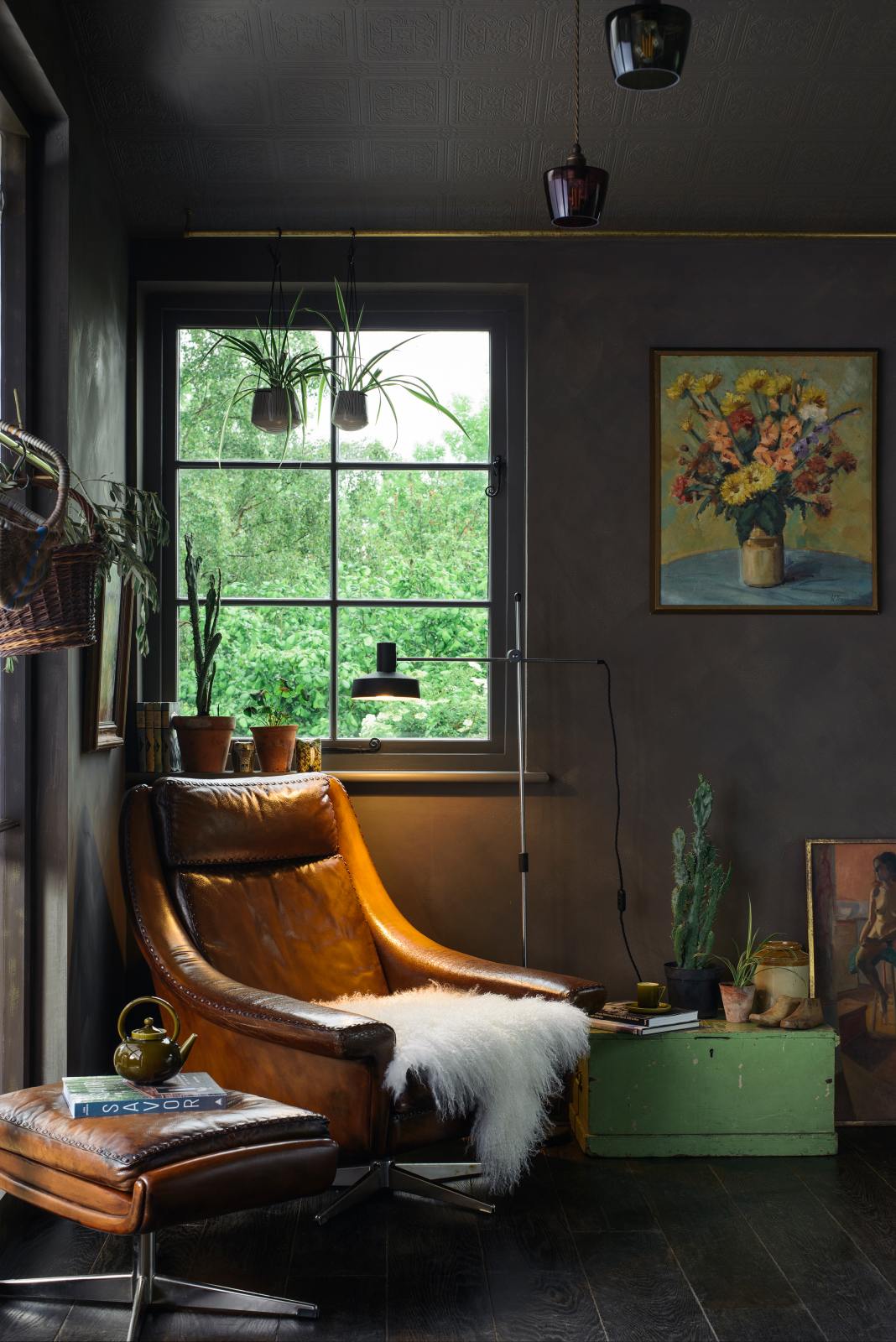 A quiet corner of a warm and inviting Haberdasher’s kitchen features a cozy reading nook. Adorned with luscious green hanging and potted plants, vintage items, a wicker basket, paintings, an artisan ceramic teapot, and a beloved armchair with a footrest, it creates a lovely spot to curl up and read a book.