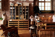 One side of a handsome range of oak furniture and lighting from the deVOL Haberdasher’s collection features a kitchen island, countertop cupboards, and copper countertop. Adorned with deVOL Heirloom accessories, Helen stools, vintage furniture and items, dressmaker mannequins, and 1900s tailor tools, this space creates a beautiful homage to the English gentlemen’s haberdashery inspiration in a bricked wall showroom.