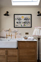 A close-up of a grand and quaint Haberdasher’s kitchen features a sink and cooker run with a Paonazzo marble worktop and decorative vanity cut, a single Belfast sink, and an aged brass Ionian tap. Beautifully decorated with tea towels, an artisan ceramic, a framed map illustration, black wall lights, a skylight, and a cloth lampshade, this kitchen creates a humble and functional space with a touch of elegance.