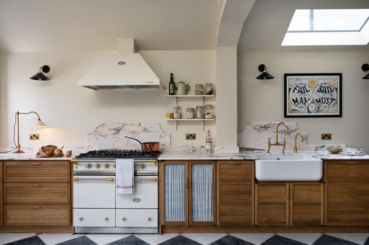 A grand and quaint Haberdasher’s kitchen features a sink and cooker run with a Paonazzo marble worktop and decorative vanity cut, a single Belfast sink, and an aged brass Ionian tap. Beautifully decorated with a white gas cooker with golden accents, a matching canopy, a deVOL grand tour desk lamp, a copper pan, artisan teapot, and dried ingredients in glass jars, this kitchen creates a humble and functional space with a touch of elegance.