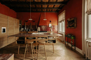 A warm and historical Haberdasher’s kitchen features a sink and cooker run with red Zellige splashback tiles, a kitchen island with an aged brass Ionian tap, a red marble worktop, and a bank of full-height cupboards. Beautifully decorated with decorative floral ceiling tiles, large narrow pleat porcelain lights, Helen stools, artisan ceramics, vintage items, and paintings, this kitchen creates a grand yet comfortable space.