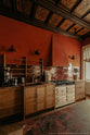 An oblique view of a warm and historical Haberdasher’s kitchen features a sink and cooker run with red Zellige splashback tiles and a kitchen island with a red marble worktop. Beautifully decorated with decorative floral ceiling tiles, artisan ceramics, vintage items, a silver coffee grinder, and a small house plant, this kitchen creates a grand yet comfortable space.