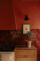 A quiet corner of a warm and historical Haberdasher’s kitchen features a sink and cooker run with red Zellige splashback tiles and a kitchen island with a red marble worktop. Beautifully decorated with a cream gas cooker, a wicker basket, a green framed illustration, and a small house plant, this kitchen creates a grand yet comfortable space.