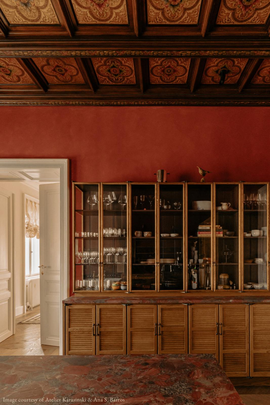 On the other side of a warm and historical Haberdasher’s kitchen features a pantry run featuring glazed countertop cupboards, a kitchen island, and a red marble worktop. Beautifully decorated with floral ceiling tiles, books, clear drinking glasses, and artisan ceramics, this kitchen creates a grand yet comfortable space.