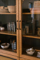 A closer look at a warm and spacious Haberdasher’s kitchen features a freestanding pantry with glazed doors. Adorned with vintage items, artisan ceramics, drinking glasses, tea towels, oxidised brass pull handles, oxidised pull hooks, and a wooden letterpress, this kitchen creates a contemporary yet nostalgic space.
