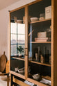 A closer look at a warm and spacious Haberdasher’s kitchen features a freestanding pantry with glazed doors. Adorned with vintage items, artisan ceramics, drinking glasses, tea towels, oxidised brass pull handles, large wooden tapas boards, and a wooden letterpress, this kitchen creates a contemporary yet nostalgic space.