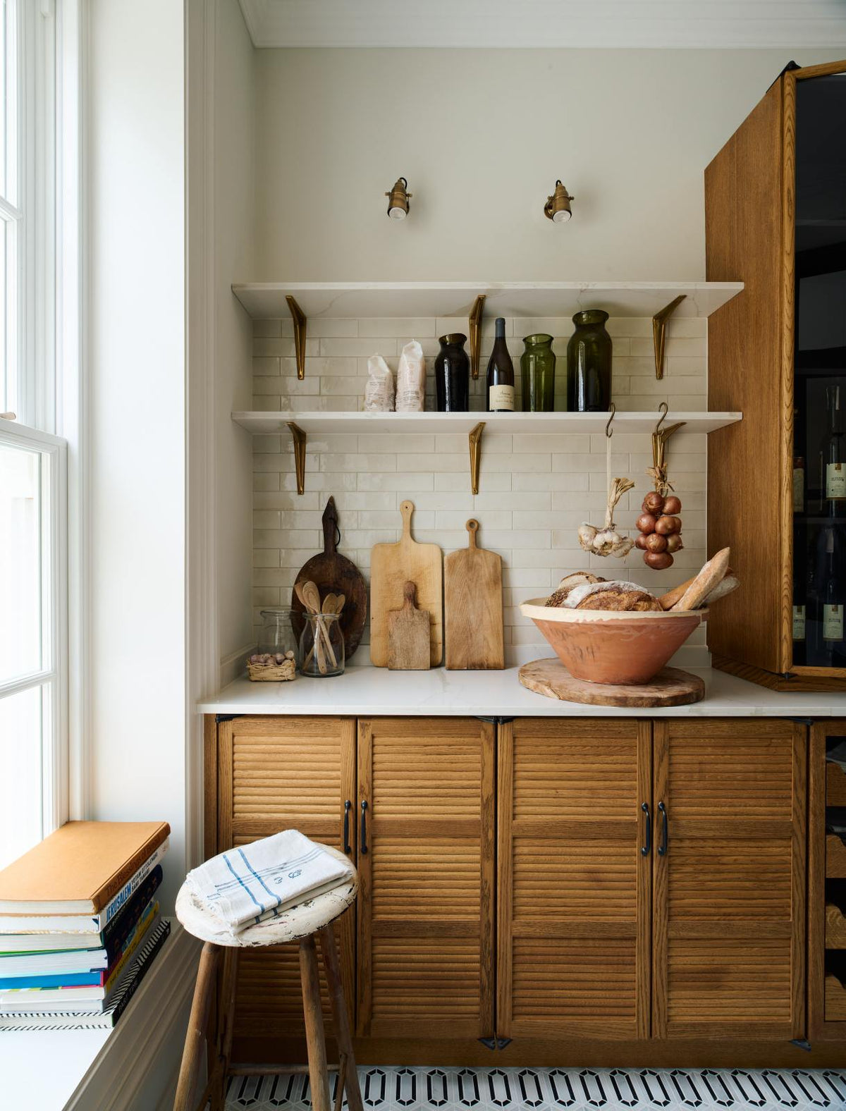 A smart and functional Haberdasher’s pantry features a long pantry run with glazed, open cupboards with internal drawers and tambour doors, a Carrara marble worktop and shelves, and cream splashback tiles. Beautifully decorated with eye-catching diamond mosaic flooring, oxidised brass hooks, cloves of garlic, onions, loaves of bread, and classic shelf brackets, this pantry creates a warm and neat storage space.