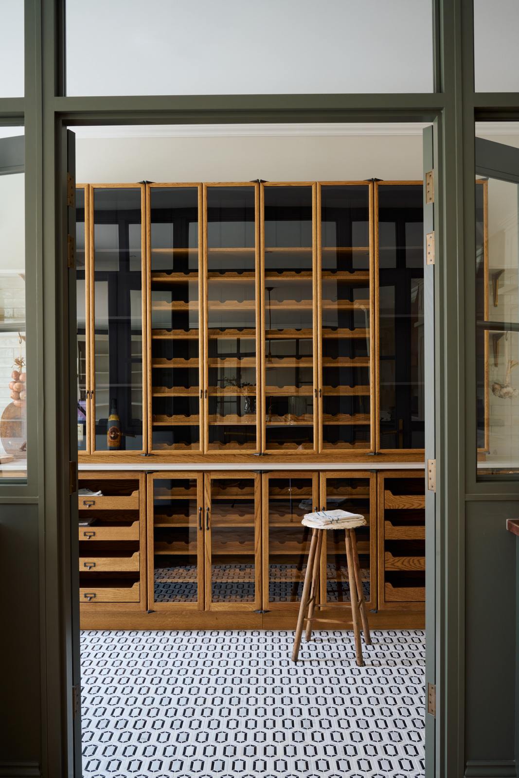 Past the olive green glass partition is a smart and functional Haberdasher’s pantry features a long pantry run with glazed, open cupboards with internal drawers and tambour doors, and a Carrara marble worktop. Beautifully decorated with eye-catching diamond mosaic flooring, vintage items, tea towels, and wine, this pantry creates a warm and neat storage space.