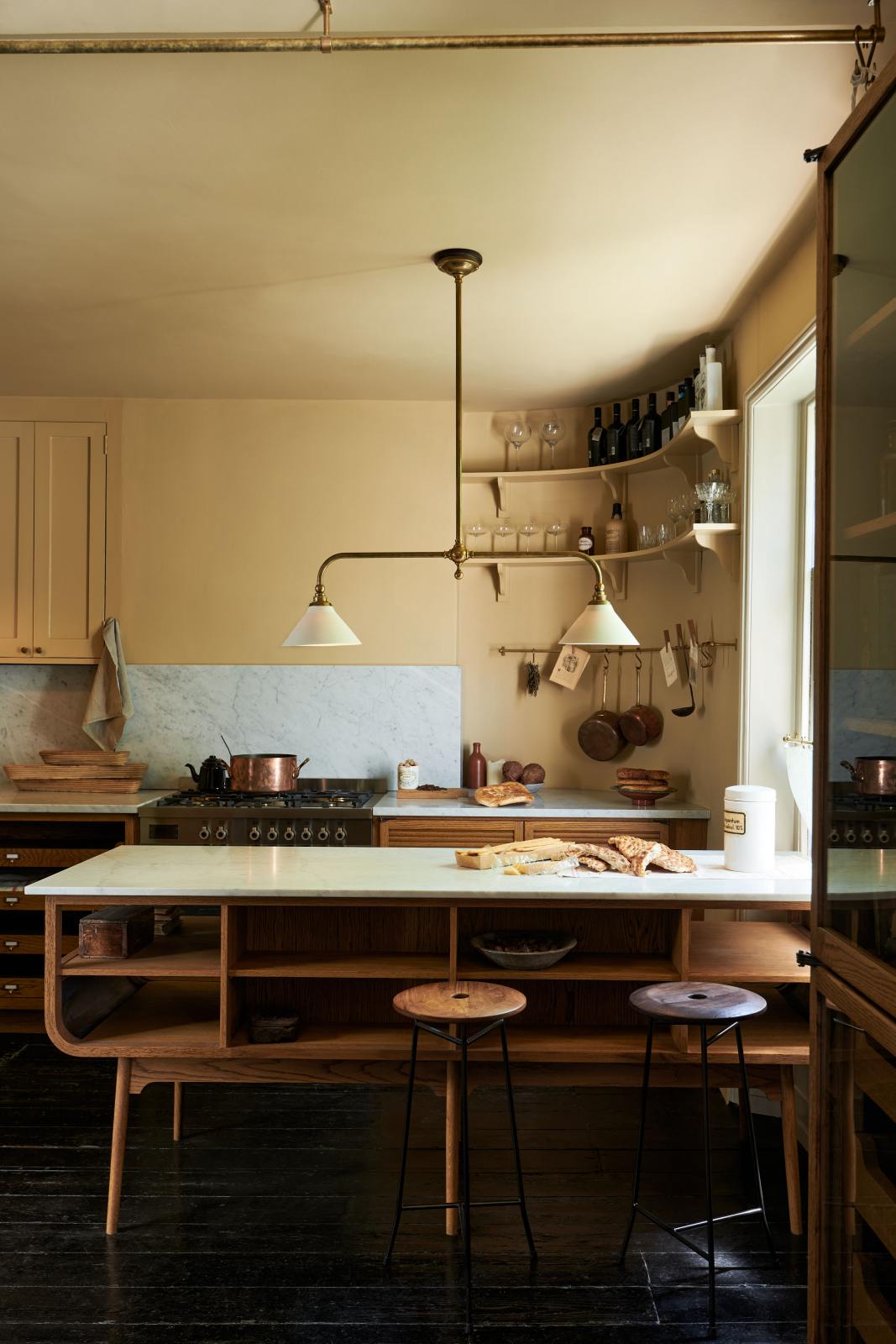 A cosy and sophisticated Haberdasher’s kitchen features a cooker run with a Carrara marble worktop, a small prep table, bespoke curved shelves, and muted yellow walls. Beautifully decorated with Creedy stools, a deVOL Heirloom gaselier light, Heirloom wall lights, aged brass hanging rails, copper pans, focaccia bread, vintage items, and artisan ceramics, this kitchen creates a bright and warm space.