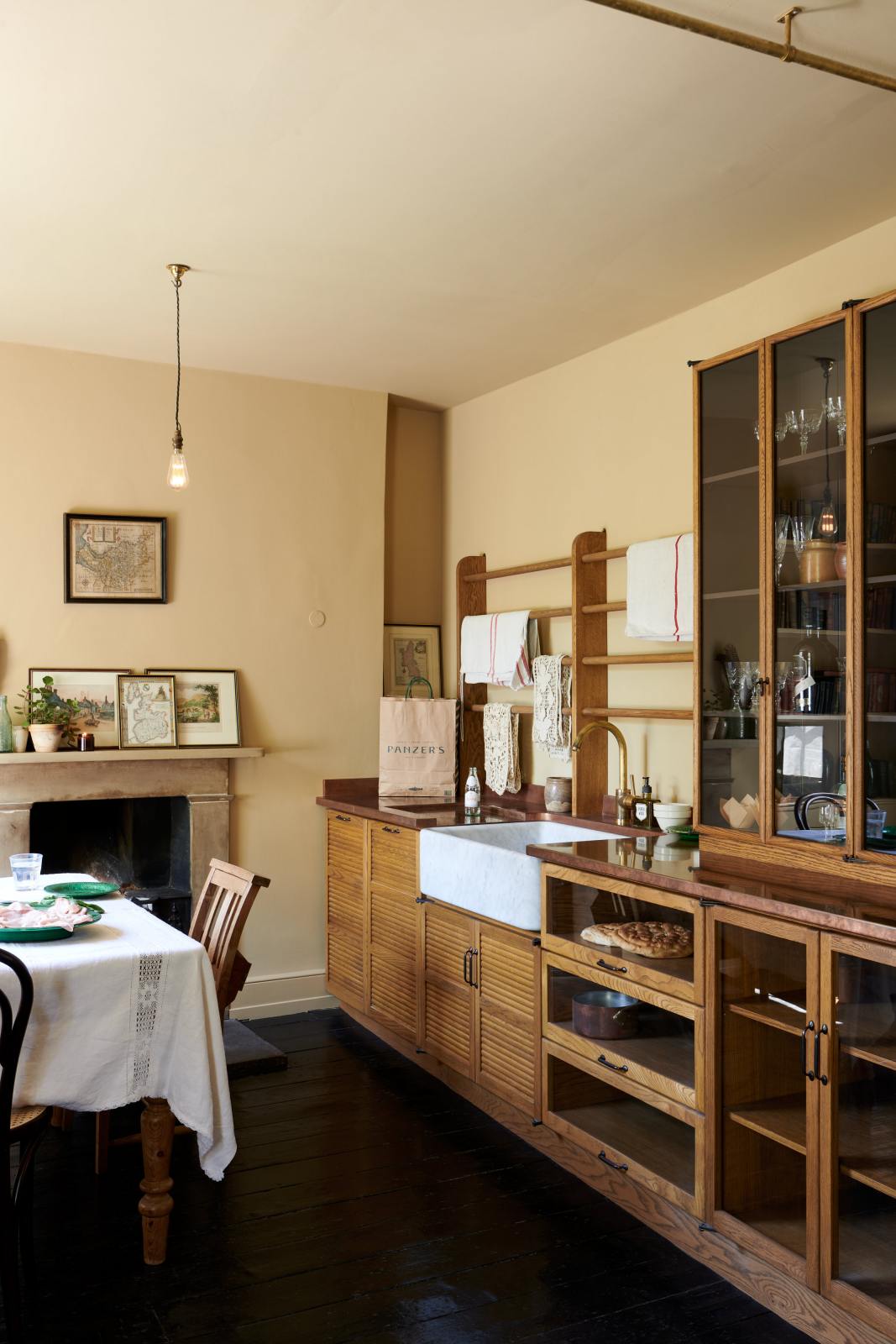 An oblique view of a cosy and sophisticated Haberdasher’s kitchen features a sink run with a copper worktop, a single marble sink, glazed countertop cupboards, and muted yellow walls. Beautifully decorated with Bentwood café chairs, vintage furniture and items, framed detailed illustrations, lace runners, tea towels, and artisan ceramics, this kitchen creates a bright and warm space.