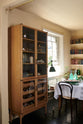 An oblique view of a cosy and sophisticated Haberdasher’s kitchen features a freestanding pantry with glazed doors. Beautifully decorated with Bentwood café chairs, vintage furniture and items, books, luscious green potted plants, aged brass café curtain rails, and artisan ceramics, this kitchen creates a bright and warm space.