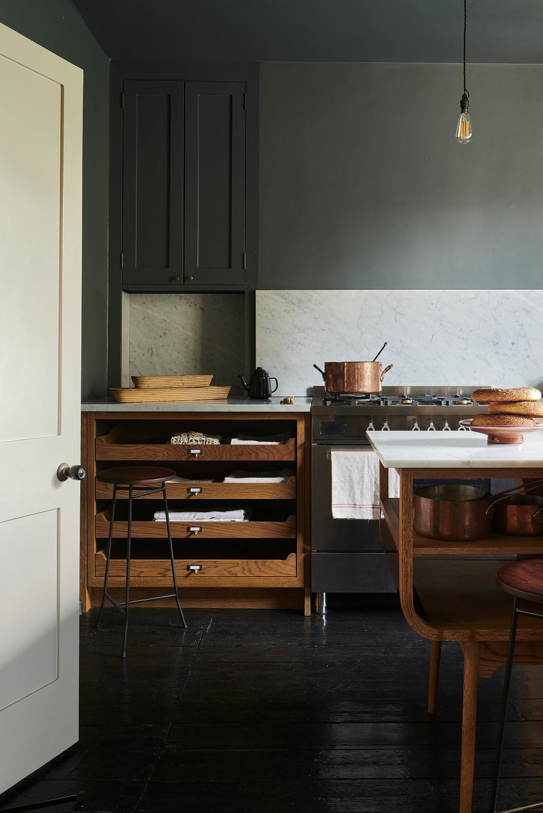 A cosy and sophisticated Haberdasher’s kitchen features a cooker run with a Carrara marble worktop, a small prep table, bespoke curved shelves, and dark charcoal walls. Beautifully decorated with stools, aged brass hanging rails, copper pans, fresh bread, vintage items, and artisan ceramics, this kitchen creates a cool yet warm and inviting space.