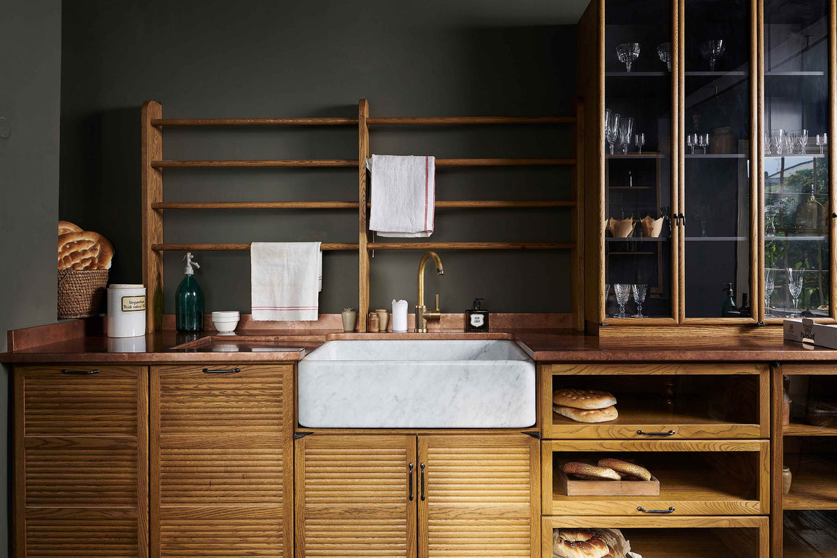 A closer look at a cosy and sophisticated Haberdasher’s kitchen features a sink run with a copper worktop, a single marble sink, glazed countertop cupboards, a freestanding pantry with glazed doors, and dark charcoal walls. Beautifully decorated with vintage furniture and items, wicker baskets, fresh bread, tea towels, and artisan ceramics, this kitchen creates a bright and warm space.