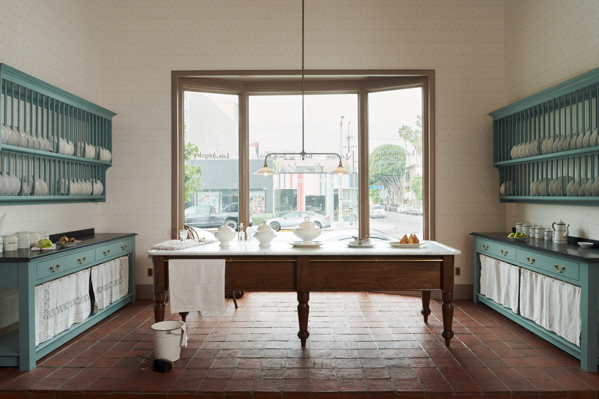 Looking out towards the large window of a beautiful and functional Trinity blue Heirloom kitchen features open cabinets, plate racks, and a showpiece Dairy table. Lovingly adorned with bone-white artisan ceramics, selected antiques, lace curtains, Gaselier lights, and earthy red terracotta square floor tiles that recreate Victorian kitchen furniture made to last a lifetime.