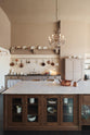 A grand Princelet pink Heirloom and Classic English kitchen features a sink cabinet with a black granite worktop, a butler’s pantry, a large kitchen island with a Carrara marble worktop, aged brass rails, ditsy delft wallpaper, a tall cream subway tile splashback, a grand tour desk lamp, and an Heirloom task light. Lovingly adorned with brass pots and pans, artisan ceramics, and an antique crystal chandelier, creating a warm, delicate, and airy kitchen space.