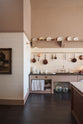 A quiet corner of a grand Princelet pink Heirloom and Classic English kitchen features a sink cabinet with a black granite worktop, a large kitchen island with a Carrara marble worktop, aged brass rails, ditsy delft wallpaper, a tall cream subway tile splashback, and an Heirloom task light. Lovingly adorned with brass pots and pans, an antique painting, and artisan ceramics, creating a warm, delicate, and airy kitchen space.