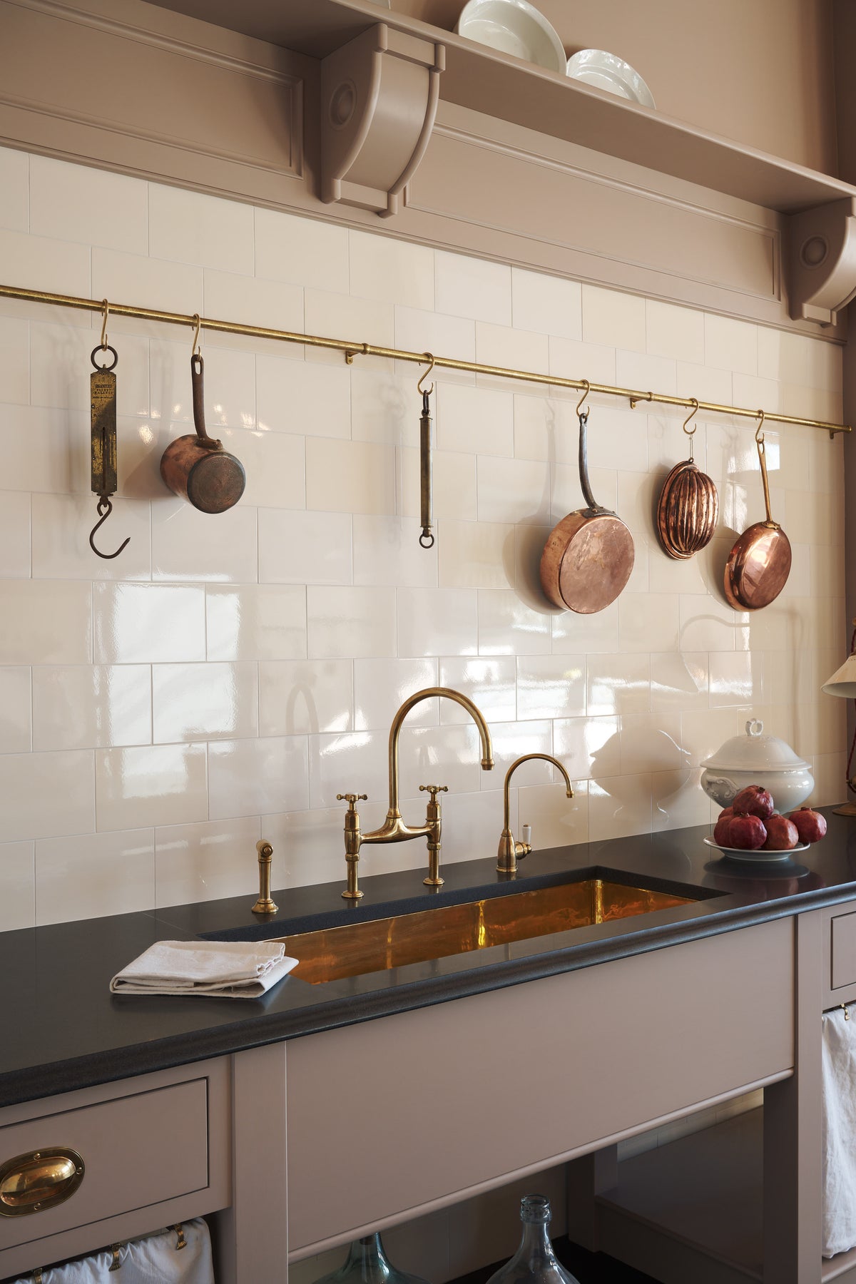 An oblique view of a quiet corner of a grand Princelet Pink Heirloom and Classic English kitchen features a sink cabinet with a black granite worktop, aged brass rails, a large polished brass sink, an aged brass Ionic tap, and a tall cream subway-tile splashback. Lovingly adorned with brass pots and pans and artisan ceramics, creating a warm, delicate, and airy kitchen space.