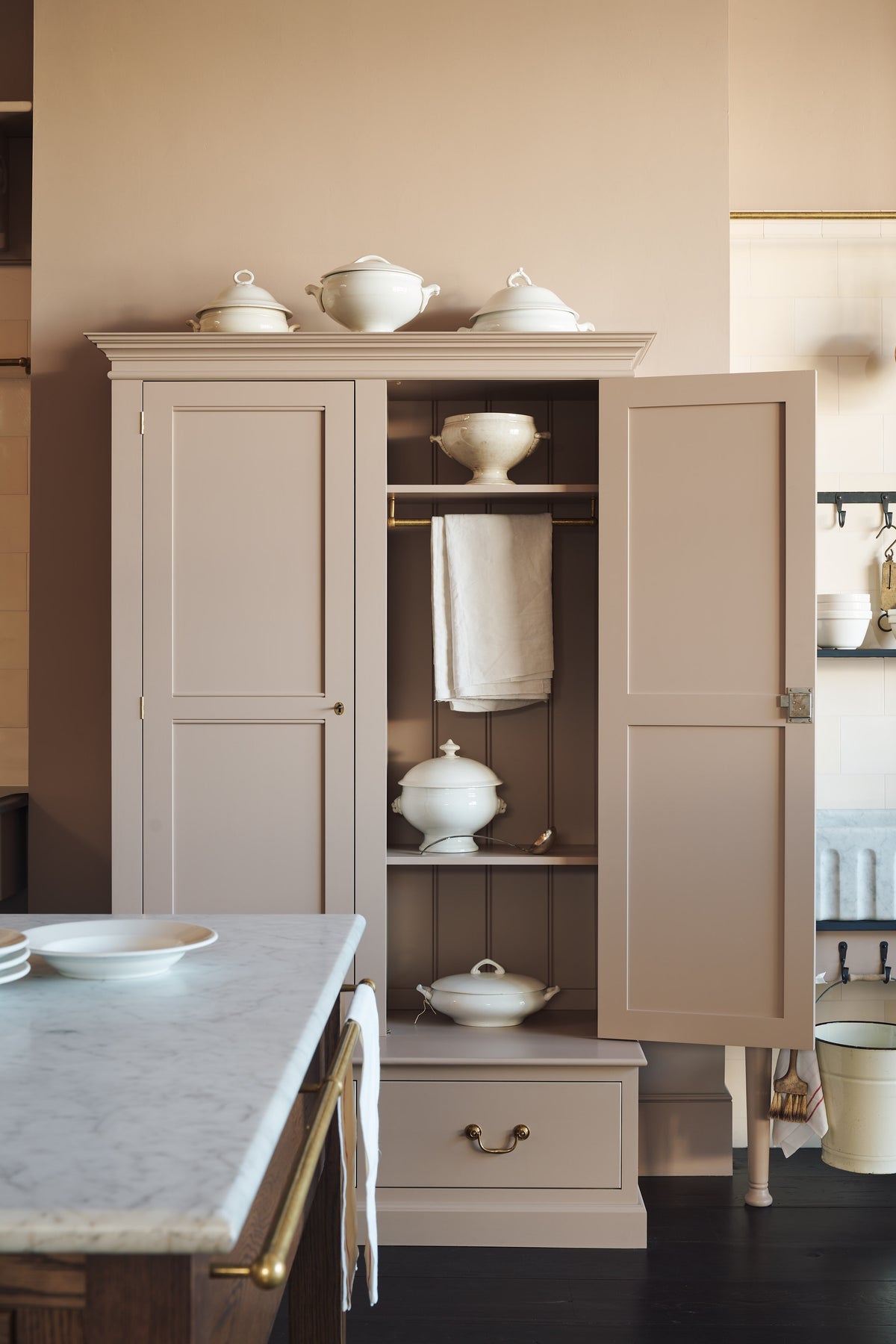 A quiet corner of a grand Princelet pink Heirloom and Classic English kitchen features a sink cabinet with a black granite worktop, a large kitchen island with a Carrara marble worktop, aged brass rails, ditsy delft wallpaper, a tall cream subway tile splashback, and an Heirloom task light. Lovingly adorned with brass pots and pans, an antique painting, and artisan ceramics, creating a warm, delicate, and airy kitchen space.