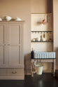 A quiet section of a grand Princelet pink Heirloom and Classic English kitchen features a Butler’s Pantry cupboard, a janitor’s sink area with a tall cream subway tile splashback and border tile, an iron hook rack, brass rails, and a fluted marble sink. Lovingly adorned with brass pots and pans and artisan ceramics, creating a warm, delicate, and airy kitchen space.