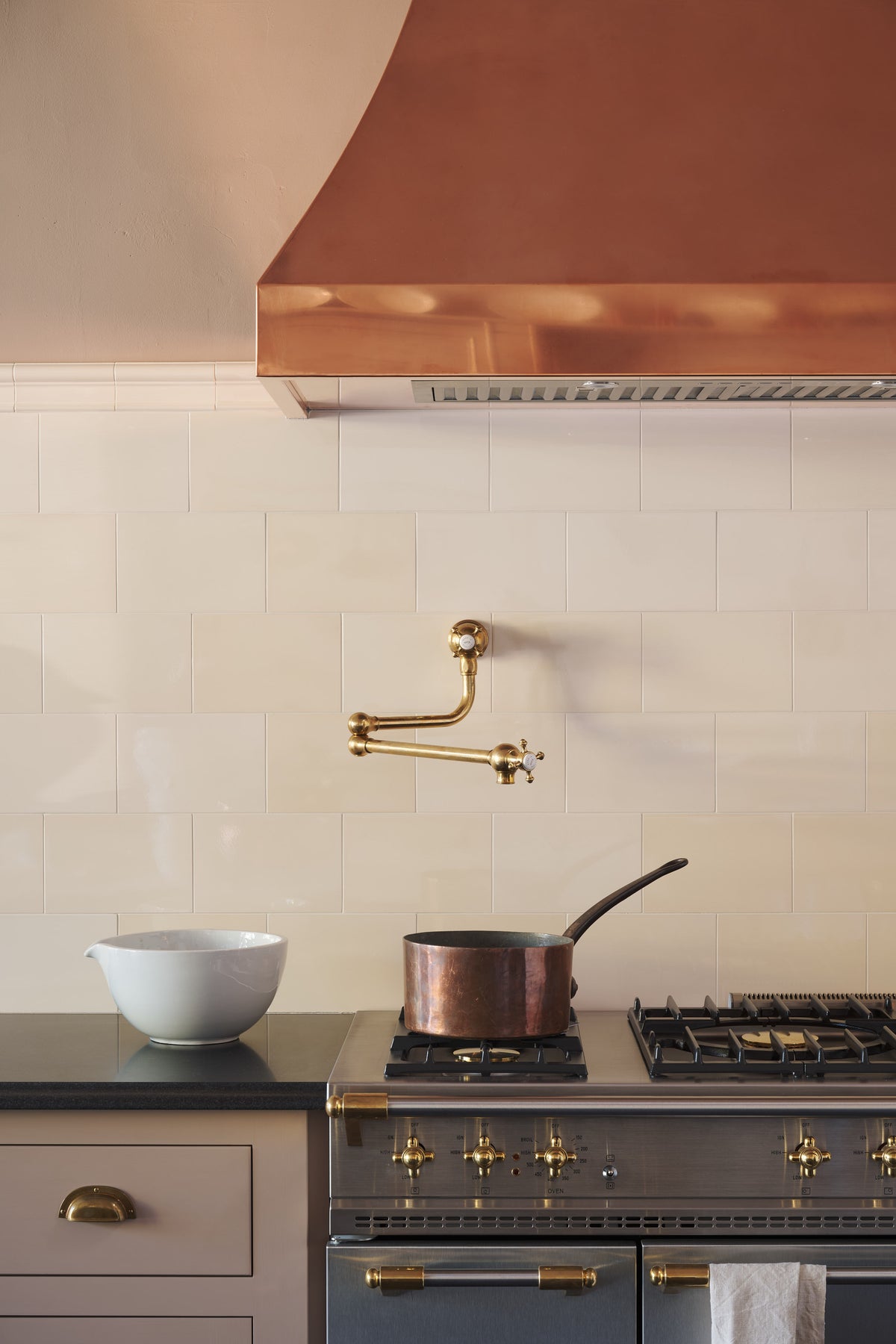 A close-up of an aged brass pot filler tap against tall cream subway tile splashback, and a large copper canopy. Adorned with a brass pan, a white ceramic bowl and a tea towel.