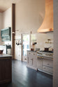 An oblique view of a quiet corner of a grand Princelet Pink Heirloom and Classic English kitchen features a cooker run with a black granite worktop, a large kitchen island with a Carrara marble worktop, an aged brass pot filler tap against a tall cream subway tile and border tile splashback, and a large copper canopy. Lovingly adorned with brass pots and pans, and artisan ceramics, creating a warm, delicate, and airy kitchen space.