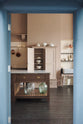 Looking through the doorway, you see a grand Princelet Pink Heirloom and Classic English kitchen. It features a Butler’s pantry cupboard, a large kitchen island with a Carrara marble worktop, and a janitor’s sink area with a tall cream subway and border-tile splashback. The space is lovingly adorned with brass pots and pans and artisan ceramics, creating a warm, delicate, and airy feel.