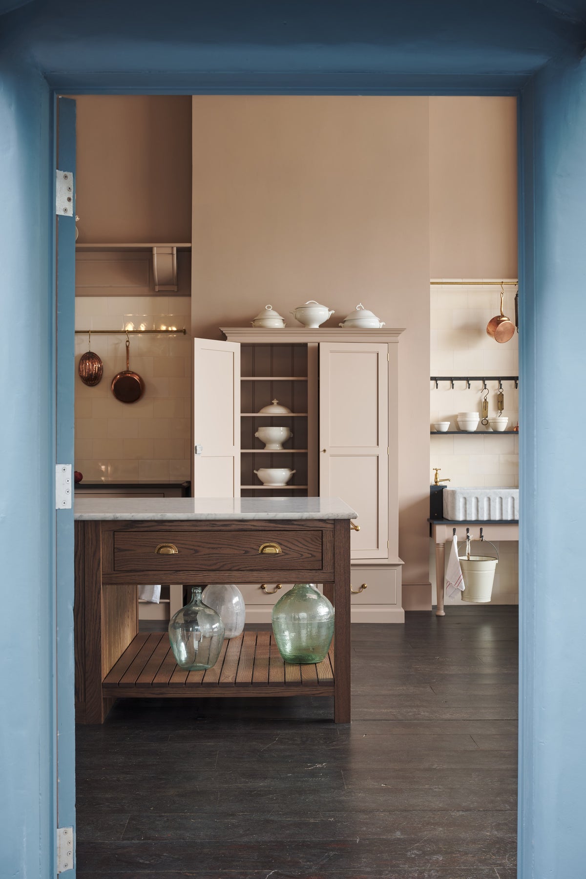 Looking through the doorway, you see a grand Princelet Pink Heirloom and Classic English kitchen. It features a Butler’s pantry cupboard, a large kitchen island with a Carrara marble worktop, and a janitor’s sink area with a tall cream subway and border-tile splashback. The space is lovingly adorned with brass pots and pans and artisan ceramics, creating a warm, delicate, and airy feel.