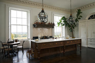 A simple yet grand Heirloom kitchen features a bespoke 3.5-metre-long dairy table with a single fluted Belfast sink, aged brass Mayan taps, a dishwasher, and a breakfast bar, creating a timeless and practical space. It is decorated with a vintage overmantle, artisan ceramics, glass and metal pendant lights, Bentwood café chairs, and a large fiddle leaf fig plant.