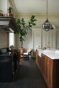 A simple yet grand Heirloom kitchen features a bespoke 3.5-metre-long dairy table with a single fluted Belfast sink, aged brass Mayan taps, a dishwasher, and a breakfast bar, creating a timeless and practical space. It is decorated with a vintage overmantle, artisan ceramics, glass and metal pendant lights, copper pans, and a large fiddle leaf fig plant.