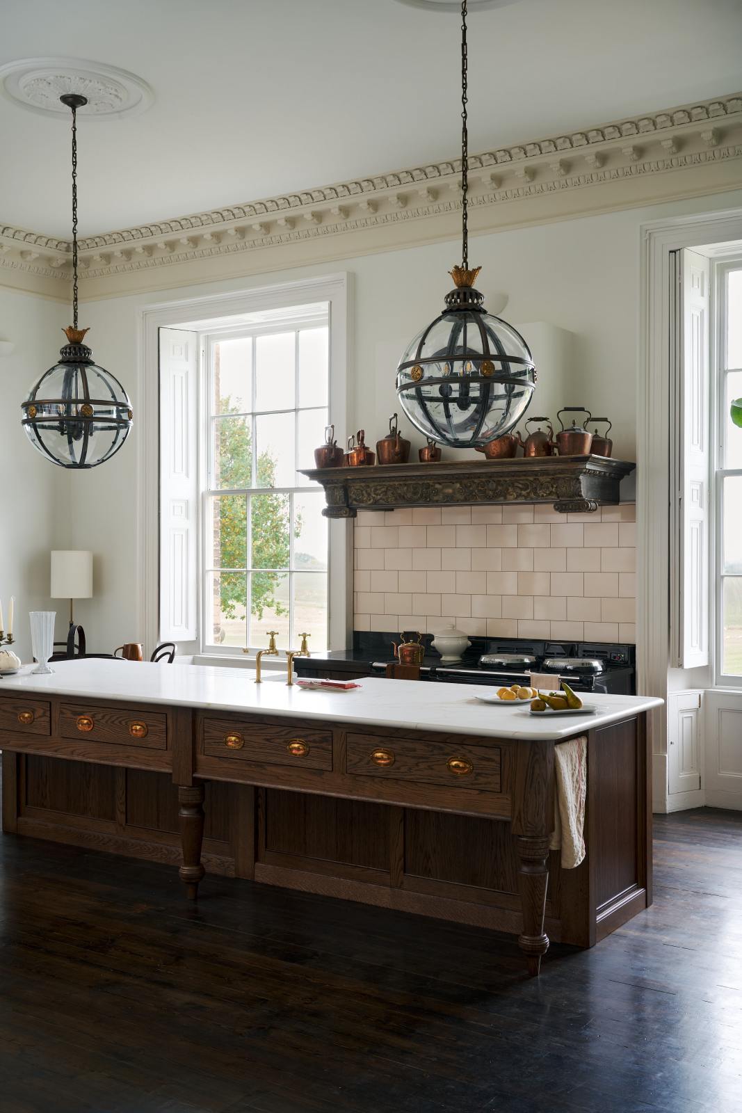 An oblique view of a simple yet grand Heirloom kitchen features a bespoke 3.5-metre-long dairy table with a single fluted Belfast sink, aged brass Mayan taps, a dishwasher, and a breakfast bar, creating a timeless and practical space. It is decorated with a vintage overmantle, artisan ceramics, glass and metal pendant lights, copper pans, and a large fiddle leaf fig plant.