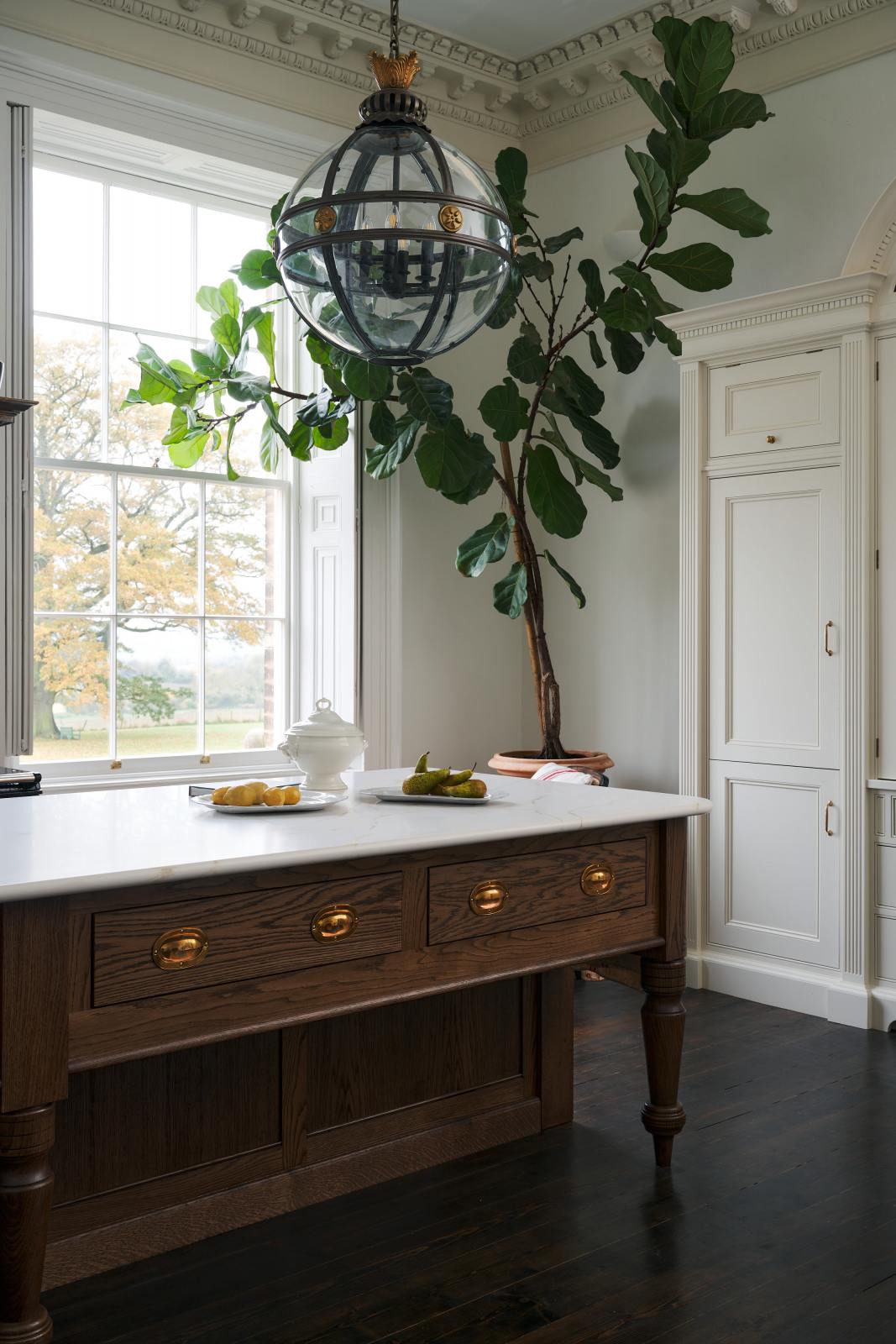 A quiet corner of a simple yet grand Heirloom kitchen features a bespoke 3.5-metre-long dairy table with a single fluted Belfast sink, aged brass Mayan taps, a dishwasher, and a breakfast bar, creating a timeless and practical space. It is decorated with a vintage overmantle, artisan ceramics, glass and metal pendant lights, Bentwood café chairs, and a golden-plated candelabra.