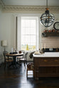 A quiet corner of a simple yet grand Heirloom kitchen features a bespoke 3.5-metre-long dairy table with a single fluted Belfast sink, aged brass Mayan taps, a dishwasher, and a breakfast bar, creating a timeless and practical space. It is decorated with a vintage overmantle, artisan ceramics, glass and metal pendant lights, Bentwood café chairs, and a golden-plated candelabra.