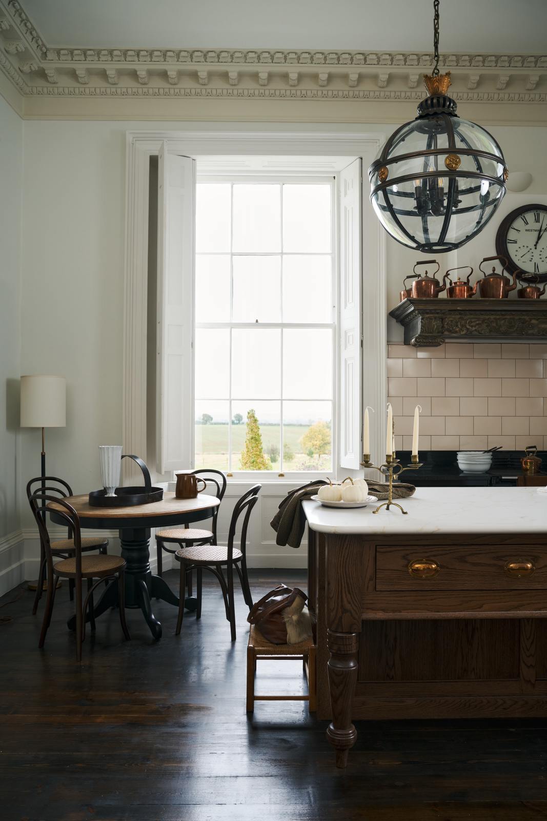 A quiet corner of a simple yet grand Heirloom kitchen features a bespoke 3.5-metre-long dairy table with a single fluted Belfast sink, aged brass Mayan taps, a dishwasher, and a breakfast bar, creating a timeless and practical space. It is decorated with a vintage overmantle, artisan ceramics, glass and metal pendant lights, Bentwood café chairs, and a golden-plated candelabra.