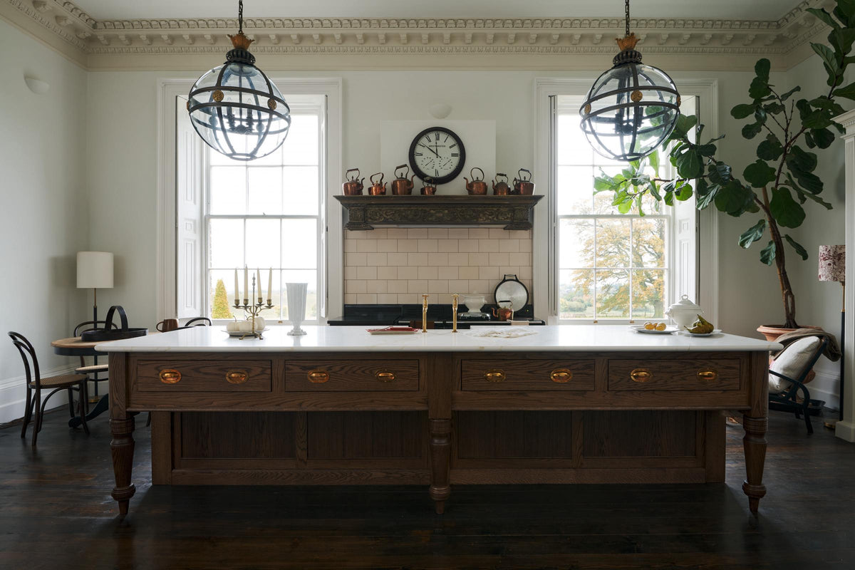 A simple yet grand Heirloom kitchen features a bespoke 3.5-metre-long dairy table with a single fluted Belfast sink, aged brass Mayan taps, a dishwasher, and a breakfast bar, creating a timeless and practical space. It is decorated with a vintage overmantle, artisan ceramics, glass and metal pendant lights, Bentwood café chairs, and a large fiddle leaf fig plant.