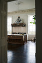 Past a white corridor is a simple yet grand Heirloom kitchen featuring a bespoke 3.5-metre-long dairy table with a single fluted Belfast sink, aged brass Mayan taps, a dishwasher, and a breakfast bar, creating a timeless and practical space. Adorned with a vintage overmantle, artisan ceramics, glass and metal pendant lights, copper pans, and a large fiddle leaf fig plant.