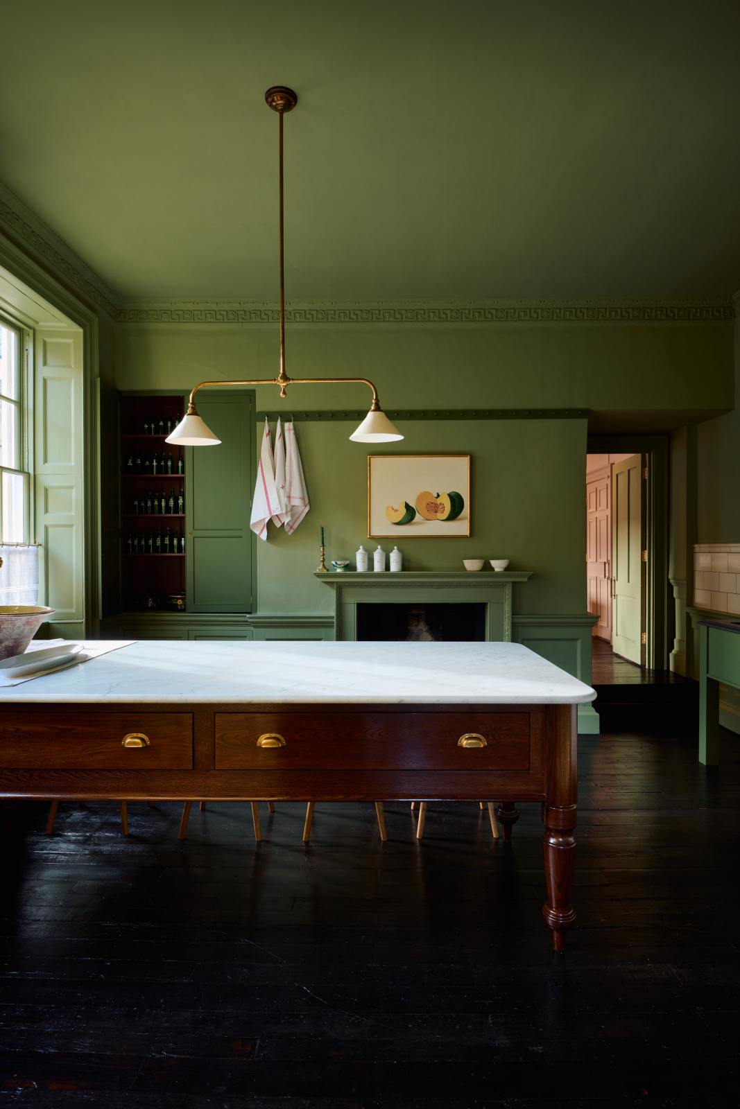 A quaint and stylish Wilkes green Heirloom kitchen features a sink cabinet, large plate rack, and dairy table. Adorned with an aged brass pot filler, artisan ceramics, Heirloom gaselier lights, cream subway splashback tiles, and a golden-framed sliced green pumpkin, this kitchen creates a luxurious dining and social space.