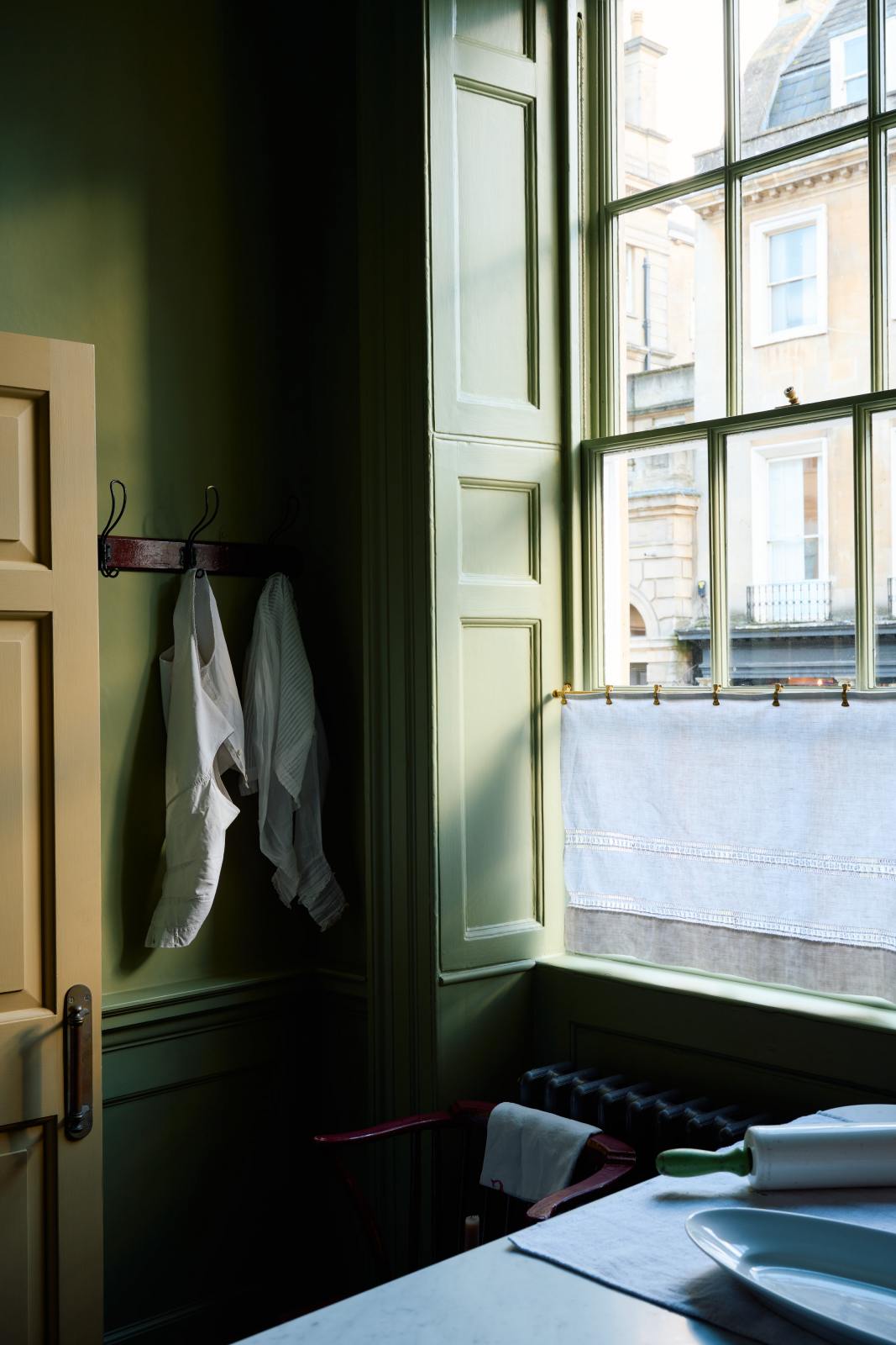A quiet corner of a quaint and stylish Wilkes green Heirloom kitchen features a large dairy table. Adorned with a café rail, a lace curtain, vintage coat hooks, and a small wooden chair, this kitchen creates a luxurious dining and social space.