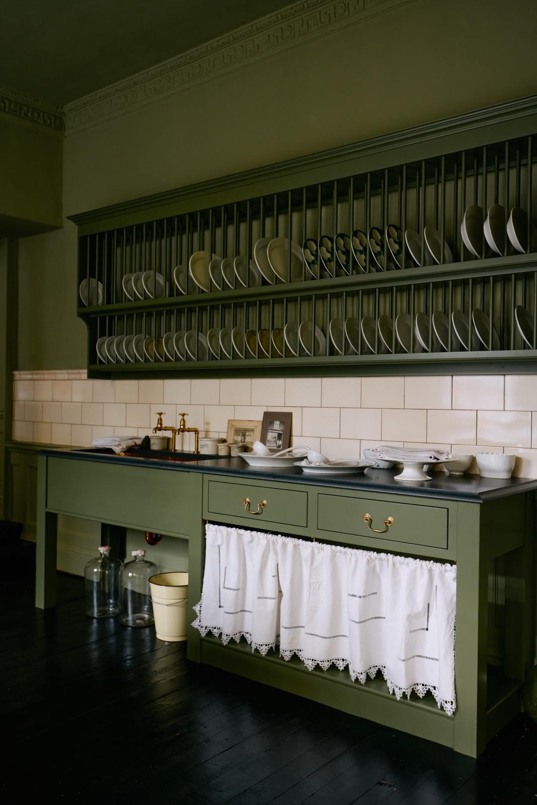 An oblique view of a quaint and stylish Wilkes green Heirloom kitchen features a sink cabinet with a café rail, white lace curtains, a polished brass sink, aged brass Mayan taps, and a large plate rack. Adorned with vintage photos, a cream bucket, simple ceramics, cream subway splashback tiles, and a golden-framed sliced green pumpkin, this kitchen creates a luxurious dining and social space.