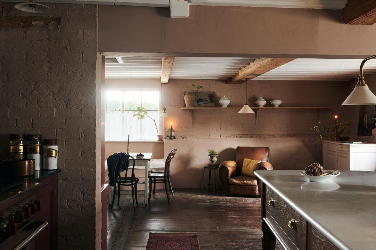 Past a refectory-red Heirloom kitchen’s cooker run and a Heirloom dairy table with an aged brass Heirloom gaselier light is a spacious dining area with vintage furniture. Beautifully decorated with a creamware pendant light, artisan ceramics, luscious green house plants, beloved candlesticks, and dried ingredients, this kitchen offers a modern twist on vintage furniture, creating a classic space designed to last.