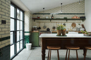 A quaint and modern Wilkes green Shaker kitchen features a cooker and sink run with a rustic oak worktop, a long shaker shelf, and a Dairy table with a breakfast nook. Adorned with a long hanging rail, signature ceramics, brass cups, and a large wall of metro tiles it embodies a playful industrial and practical design. 
