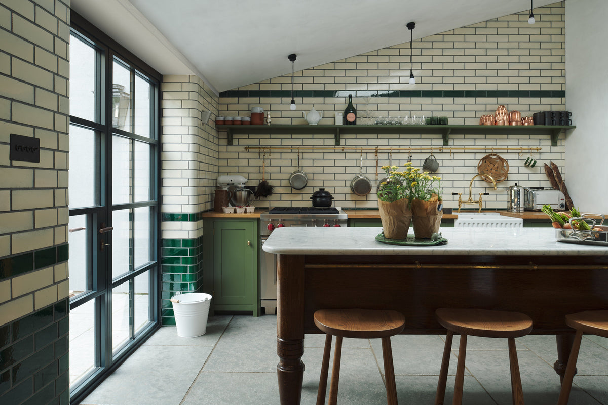 A quaint and modern Wilkes green Shaker kitchen features a cooker and sink run with a rustic oak worktop, a long shaker shelf, and a Dairy table with a breakfast nook. Adorned with a long hanging rail, signature ceramics, brass cups, and a large wall of metro tiles it embodies a playful industrial and practical design. 