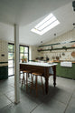 An oblique view of a quaint and modern Wilkes green Shaker kitchen features a cooker and sink run with a rustic oak worktop, a long shaker shelf, and a Dairy table with a breakfast nook. Adorned with a long hanging rail, sky light, signature ceramics, brass cups, and a large wall of metro tiles it embodies a playful industrial and practical design.