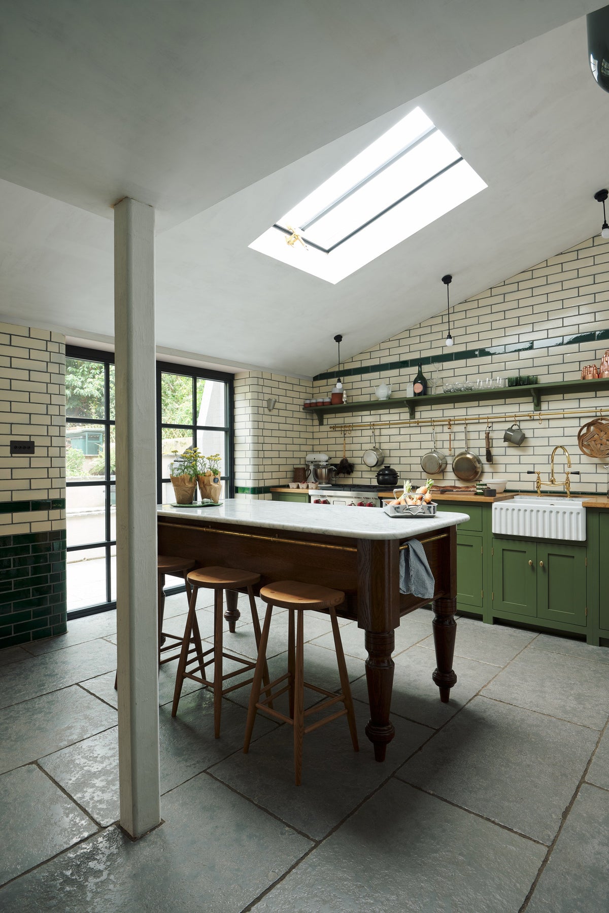 An oblique view of a quaint and modern Wilkes green Shaker kitchen features a cooker and sink run with a rustic oak worktop, a long shaker shelf, and a Dairy table with a breakfast nook. Adorned with a long hanging rail, sky light, signature ceramics, brass cups, and a large wall of metro tiles it embodies a playful industrial and practical design.