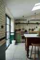 A quaint and modern Wilkes green Shaker kitchen features a cooker and sink run with a rustic oak worktop, a long shaker shelf, and a Dairy table with a breakfast nook. Adorned with a long hanging rail, signature ceramics, brass cups, and a large wall of metro tiles it embodies a playful industrial and practical design.