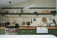 A closer look at the quaint and modern Wilkes green Shaker kitchen features a cooker and sink run with a rustic oak worktop, a long shaker shelf, and a Dairy table with a breakfast nook. Adorned with a long hanging rail, signature ceramics, brass cups, and a large wall of metro tiles it embodies a playful industrial and practical design.