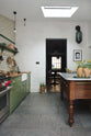Looking past the quaint and modern Wilkes green Shaker kitchen features a cooker and sink run with a rustic oak worktop, a long shaker shelf, and a Dairy table with a breakfast nook is an open doorway to the living space. Adorned with a long hanging rail, signature ceramics, brass cups, and a large wall of metro tiles it embodies a playful industrial and practical design.